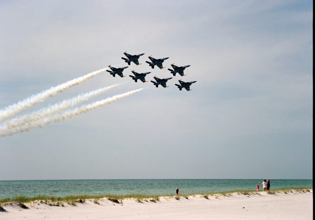 Six U.S. Navy Blue Angels F/A-18 Super Hornets in tight diamond formation over a sugar-white sand Gulf of Mexico beach with families on the sand watching.