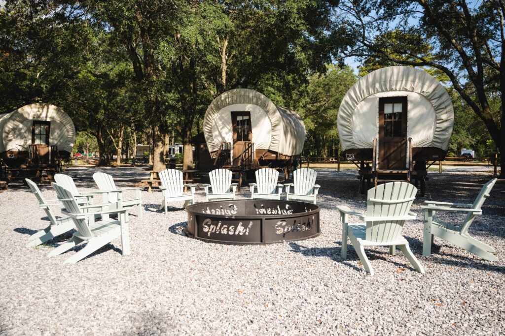 Three covered Conestoga glamping wagons and a Splash fire pit ringed by Adirondack chairs under mature trees at Splash RV Resort.