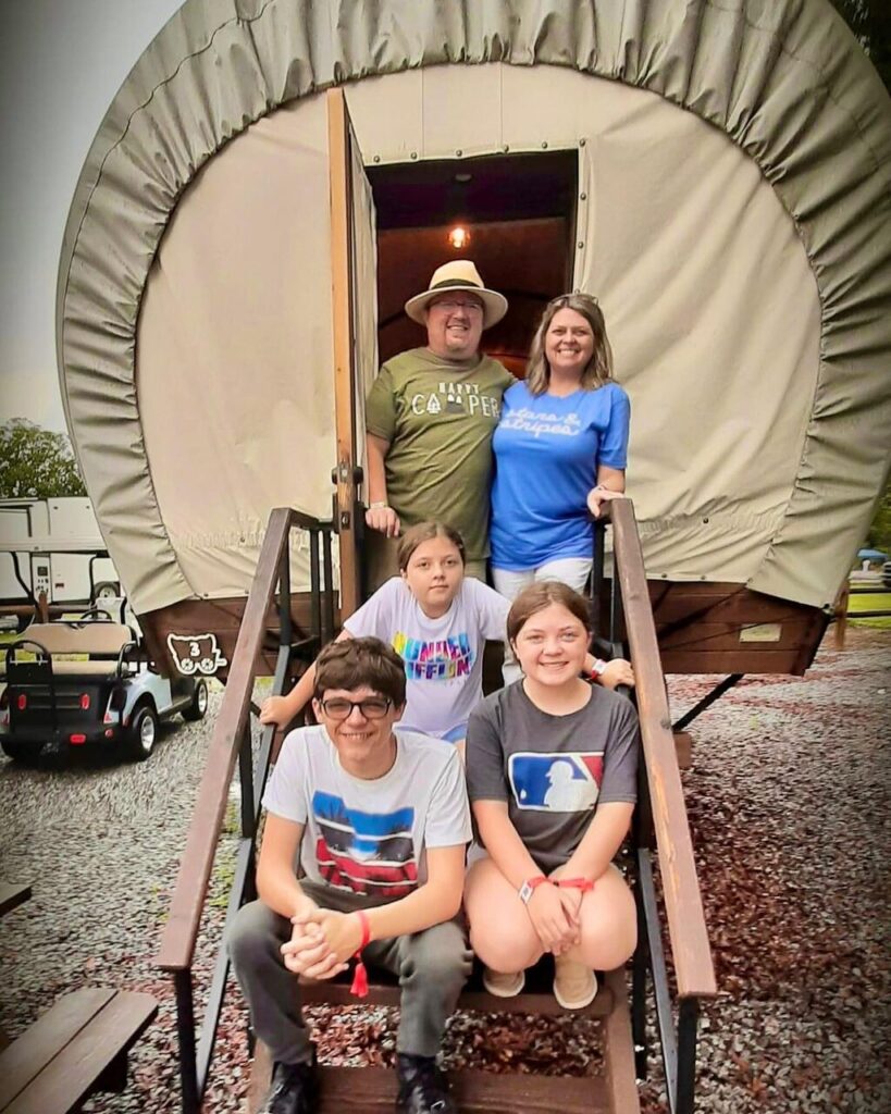 Family on the steps of a Conestoga-style glamping wagon at Splash RV Resort