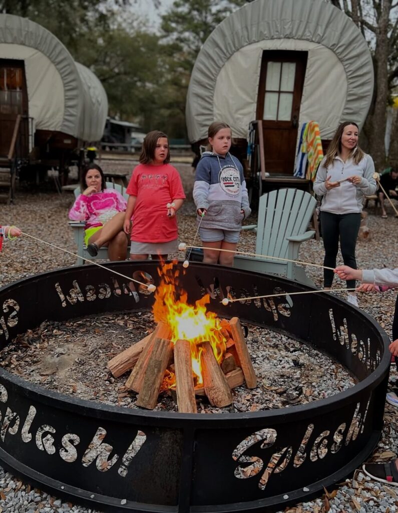 Family roasting marshmallows near Conestoga glamping wagons at Splash RV Resort