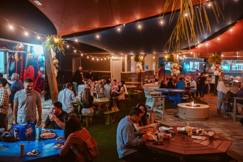 Guests dining at picnic tables beneath string lights and shade sails on an evening at Splash RV Resort, a short drive from Pensacola Beach