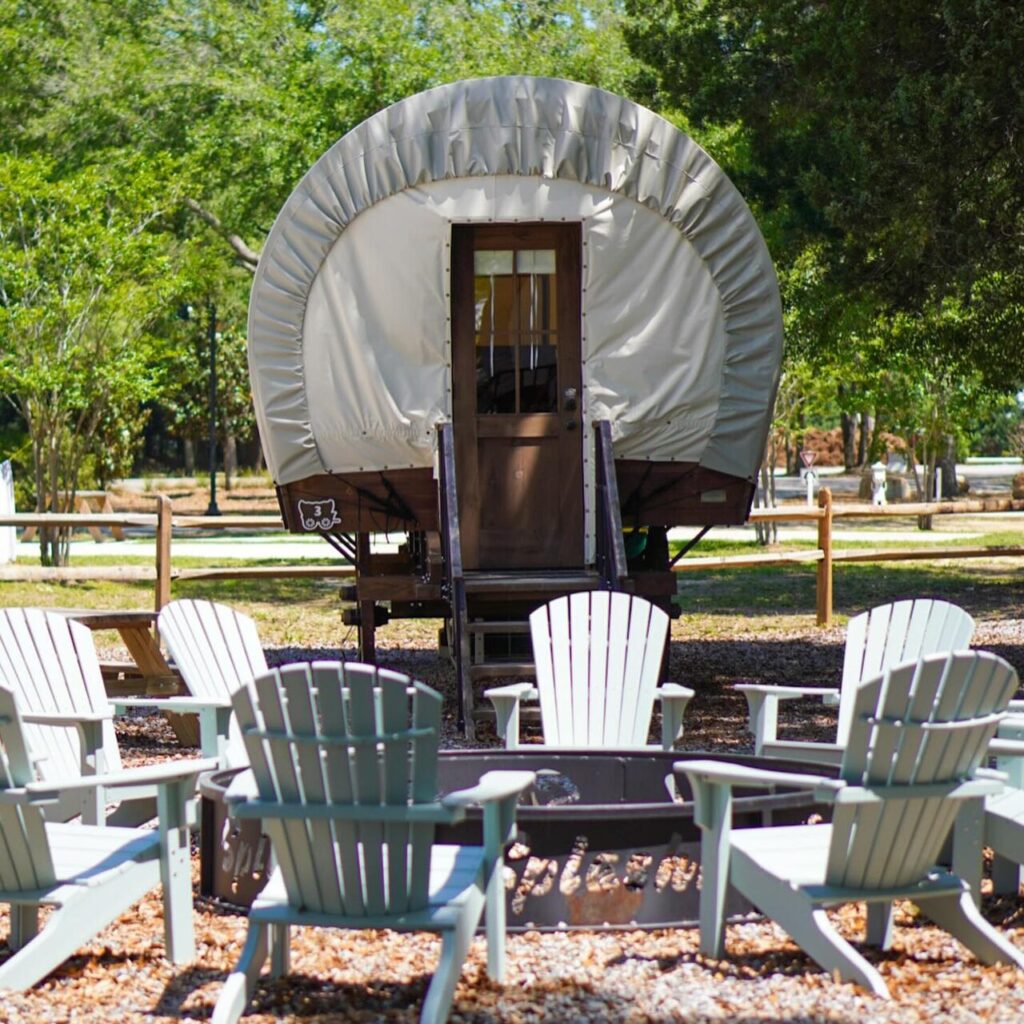 A Conestoga style glamping wagon with Adirondack chairs circled around a fire ring at an RV resort near Pensacola Beach, Florida