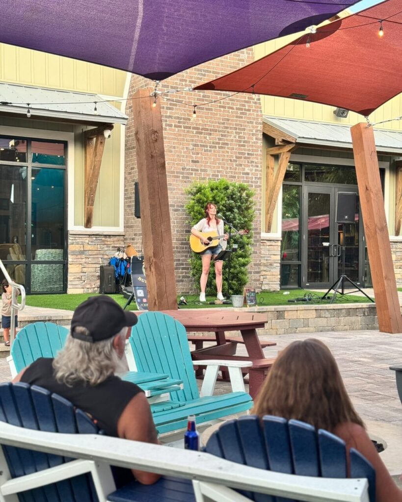 Guests relaxing in Adirondack chairs watching live acoustic music under shade sails at an RV resort near Pensacola Beach, Florida