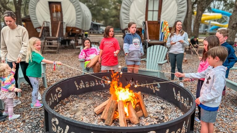 Families roasting marshmallows around a campfire ring beside Conestoga glamping wagons at Splash RV Resort near Pensacola Beach
