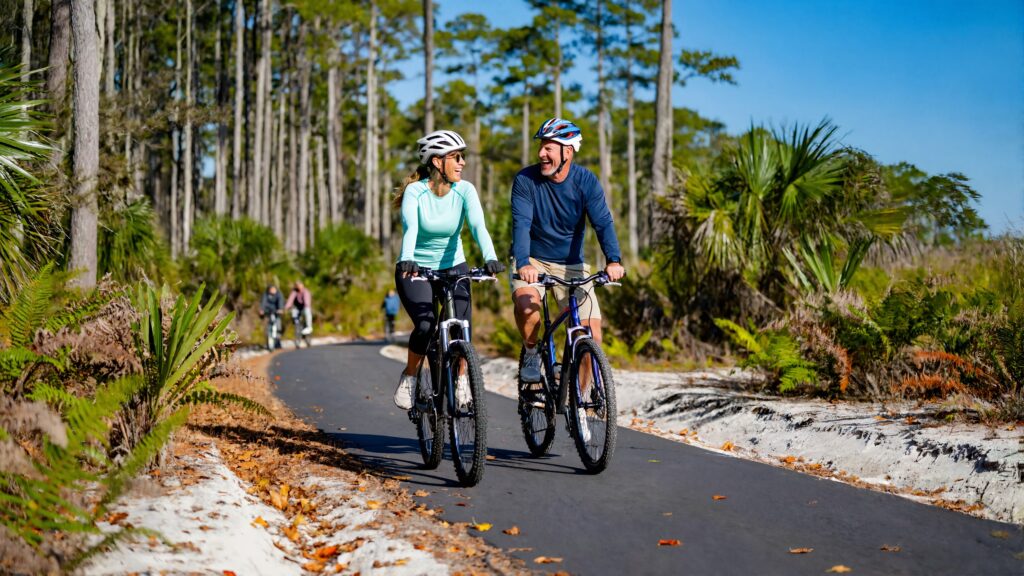A man and a woman in helmets ride bicycles side by side on a paved trail lined with pines and palmettos on a sunny fall morning