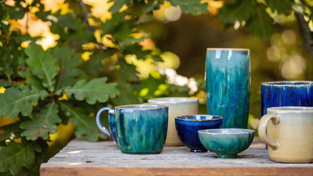 Handmade glazed ceramic mugs, bowls and a tall vase in teal, cobalt and cream on a weathered wooden table outdoors under oak leaves
