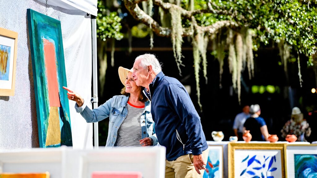 A smiling woman in a straw hat points out a colorful abstract painting to a man at an outdoor art festival booth under moss-draped oak trees