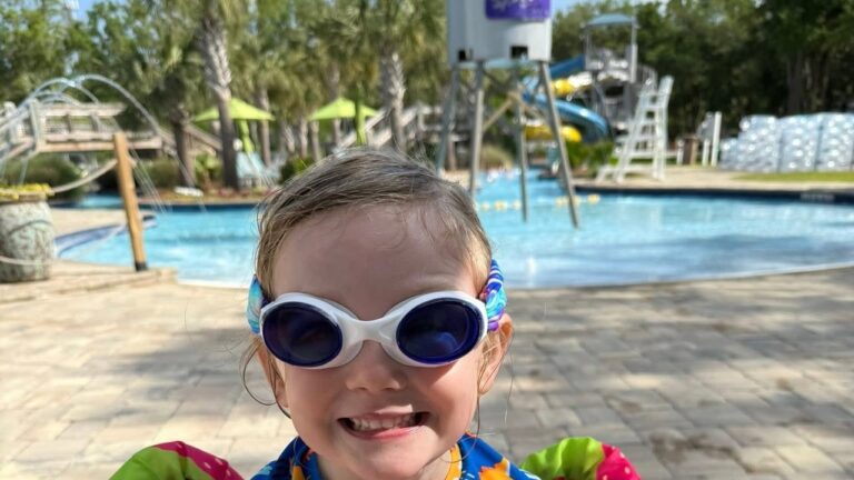 A smiling girl in goggles and a dinosaur rash guard on the pool deck at Splash RV Resort near Navarre Beach, with the pool and water slides behind her