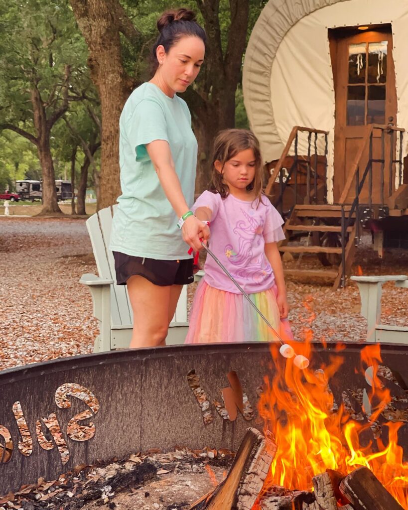 A woman and a young girl roasting marshmallows at a fire ring in front of a Conestoga glamping wagon at Splash RV Resort near Pensacola