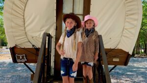 Two girls in cowboy hats and boots on the steps of a Conestoga glamping wagon at Splash RV Resort, a home base for things to do in Fort Walton Beach