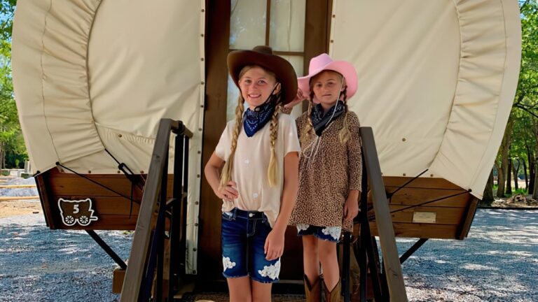 Two girls in cowboy hats and boots on the steps of a Conestoga glamping wagon at Splash RV Resort, a home base for things to do in Fort Walton Beach
