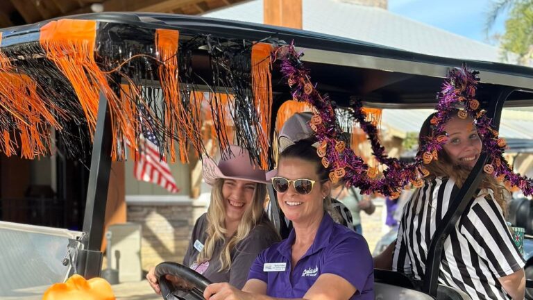 Three smiling women ride a golf cart decorated with orange and purple Halloween garland at Splash RV Resort in Milton, FL