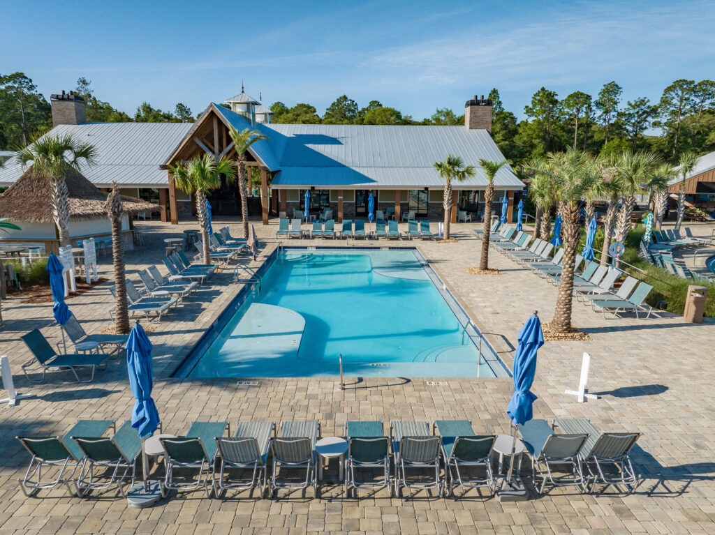 Rows of lounge chairs and blue umbrellas around a heated pool at Splash RV Resort near Pensacola, with a metal-roofed building and palm trees behind