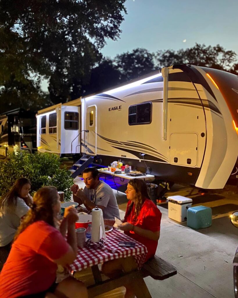 Four guests eating dinner at a checkered picnic table beside an RV with LED trim lights on a concrete site at dusk, at a luxury RV resort in Florida near Navarre Beach