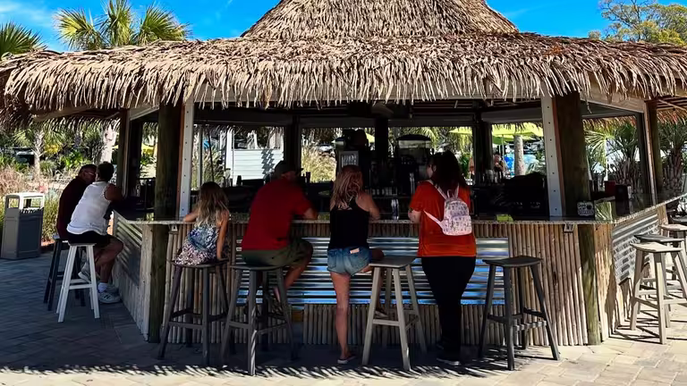 Guests seated on stools at the thatched-roof tiki bar at Splash RV Resort, a luxury RV resort in Florida near Pensacola