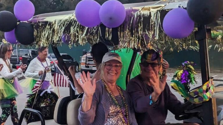 Smiling couple in Mardi Gras beads waving from a golf cart decorated with purple and black balloons and gold tinsel at Splash RV Resort near Pensacola