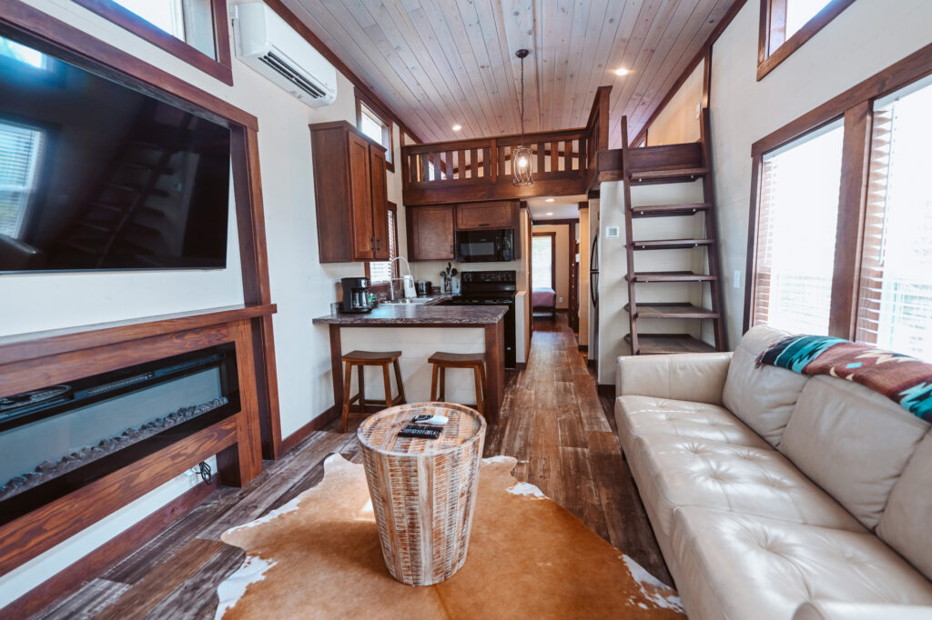 Inside a monthly rental cottage at Splash RV Resort near Pensacola, with a cream leather sofa, a wall TV over an electric fireplace, a kitchen bar with two stools and stairs up to the sleeping loft