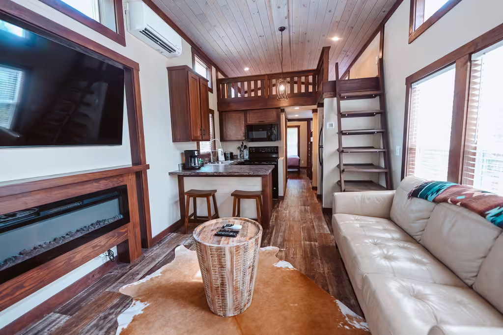 Inside a monthly rental cottage at Splash RV Resort near Pensacola, with a cream leather sofa, a wall TV over an electric fireplace, a kitchen bar with two stools and stairs up to the sleeping loft