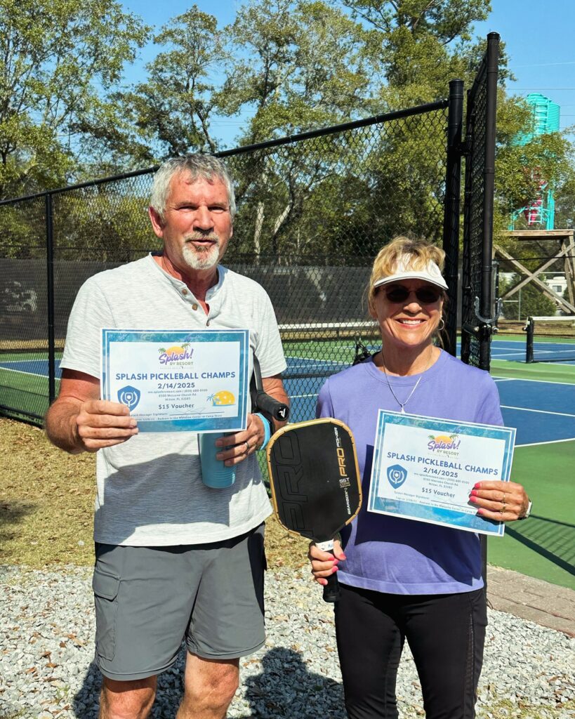 A smiling man and woman holding Splash Pickleball Champs certificates and a paddle beside the fenced pickleball courts at Splash RV Resort near Navarre Beach