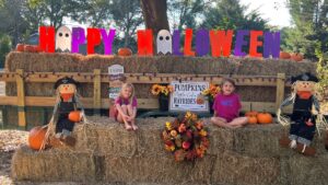Two girls sitting on hay bales beside a Happy Halloween display with scarecrows and pumpkins at Splash RV Resort near Pensacola