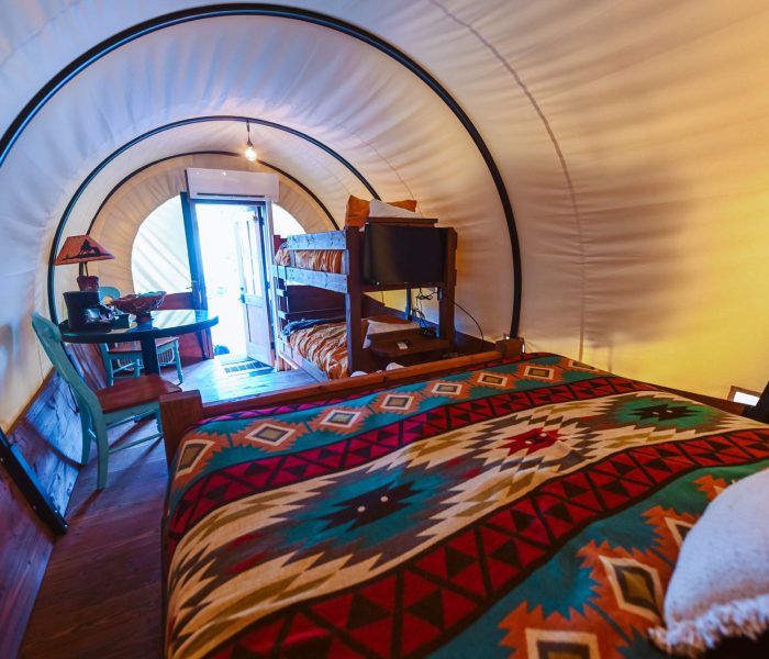 The interior of a Conestoga-style glamping wagon at Splash RV Resort in Milton, Florida features a plush bed dressed in a vibrant Southwestern-patterned blanket of teal, red, and orange, alongside wooden bunk beds, a flat-screen TV, and a quaint bistro table with a turquoise chair beneath the curved white canvas canopy. Warm golden lighting illuminates the rich hardwood floors and cozy furnishings, creating an inviting, adventurous atmosphere that blends frontier charm with modern luxury.
