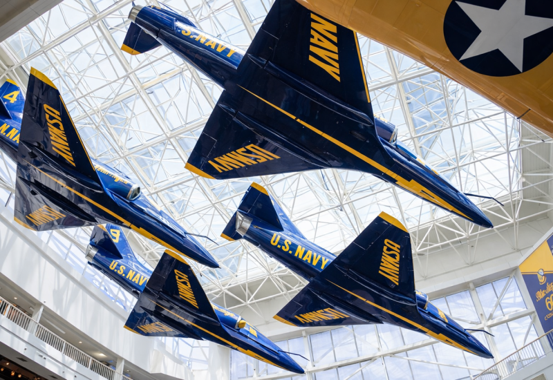 Four blue and yellow U.S. Navy Blue Angels jets suspended in diamond formation from the ceiling of a museum atrium with glass-paneled geometric roof and natural light at Splash RV Resort in Milton, Florida.