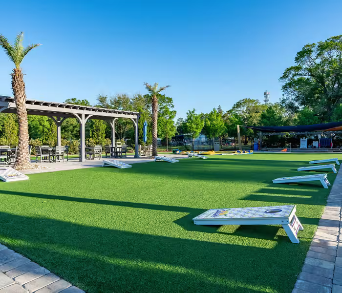 Cornhole boards on bright green turf at Splash RV Resort, bordered by a pergola and palm trees.