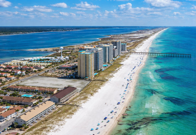 This aerial photo captures a stunning Florida coastline featuring a narrow barrier island lined with high-rise beachfront condominiums, white sandy beaches dotted with colorful umbrellas, and a long fishing pier extending into turquoise Gulf waters on the right, with a calm bay on the left. The bright blue sky scattered with puffy white clouds and the vivid gradient of emerald-to-deep-blue ocean create a vibrant, sunny resort atmosphere.