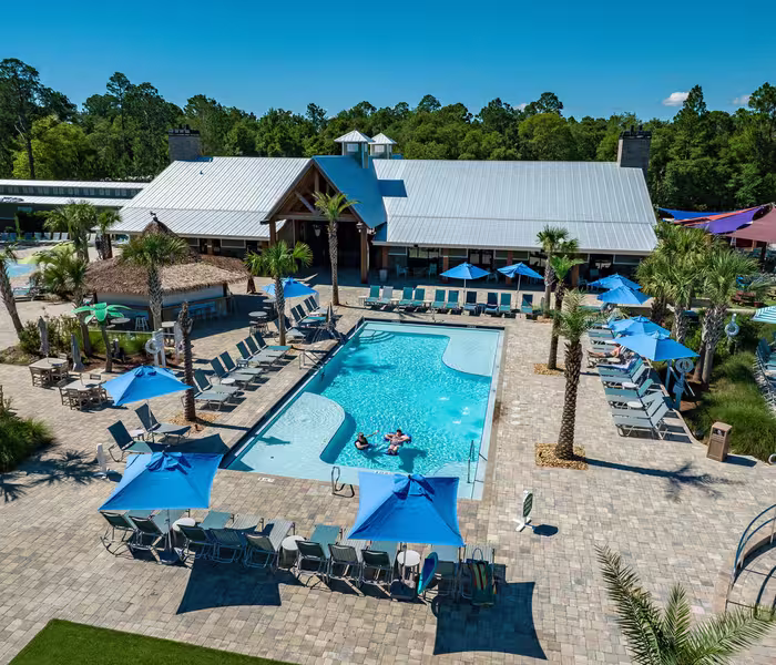 Aerial view of resort-style pool with turquoise water, blue umbrellas, tiki bar, and clubhouse at Splash RV Resort in Milton, Florida.