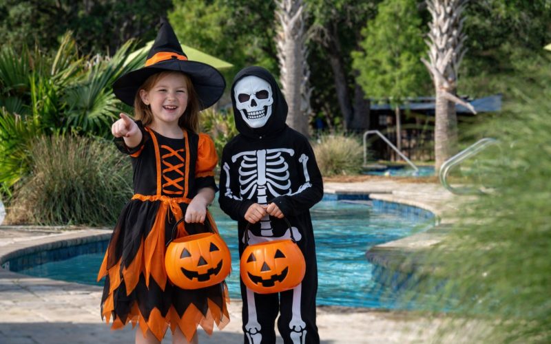 Two children in Halloween costumes holding pumpkin buckets by the swimming pool at Splash RV Resort.