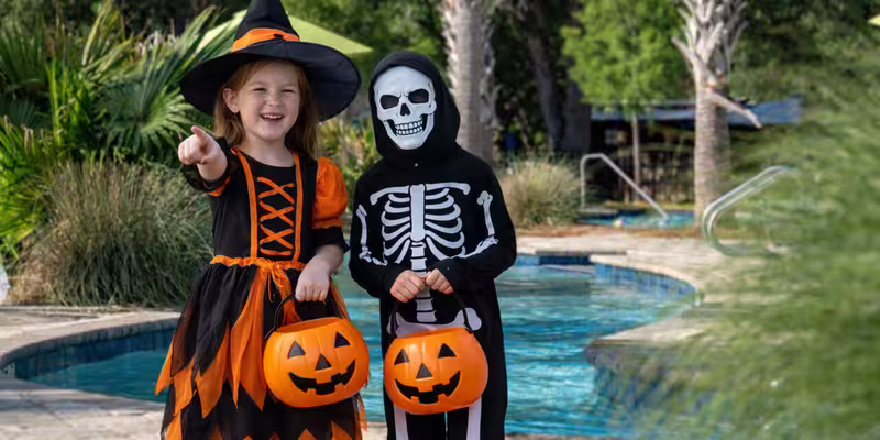 Two children in Halloween costumes holding pumpkin buckets by the swimming pool at Splash RV Resort.