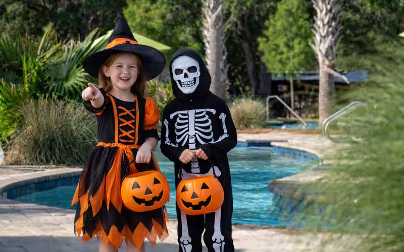 Two children in Halloween costumes holding pumpkin buckets by the swimming pool at Splash RV Resort.