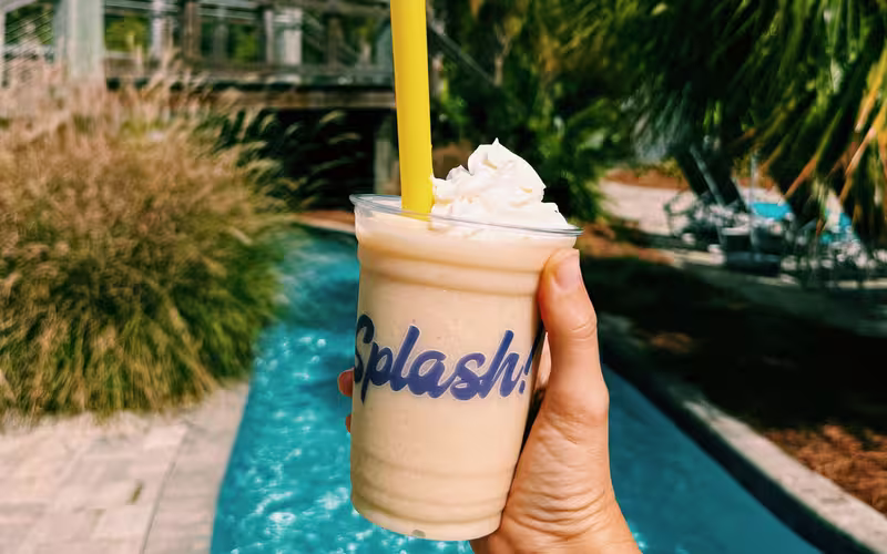Hand holding a frozen drink with the Splash RV Resort logo in front of the resort's winding lazy river.