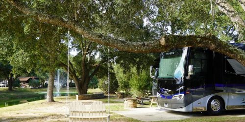 Luxury motorhome parked under a large oak tree at Splash RV Resort with a white porch swing and pond fountain in the background.