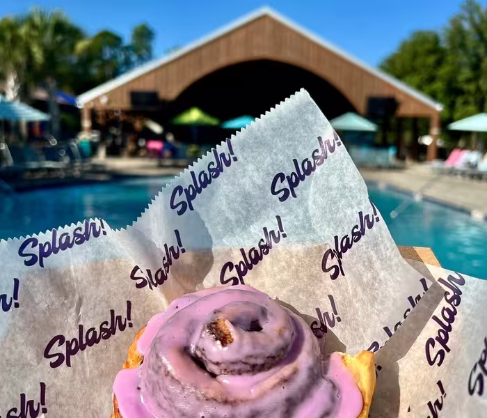 Pink frosted cinnamon roll in branded paper with a swimming pool and wooden pavilion in the background at Splash RV Resort