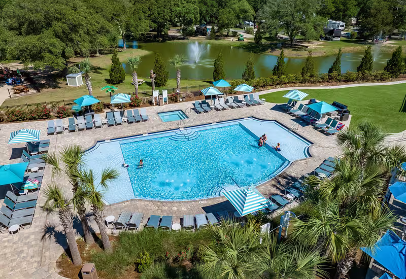 Aerial view of Splash RV Resort sparkling pool with striped umbrellas lounge chairs and palm trees with lake and fountains in background