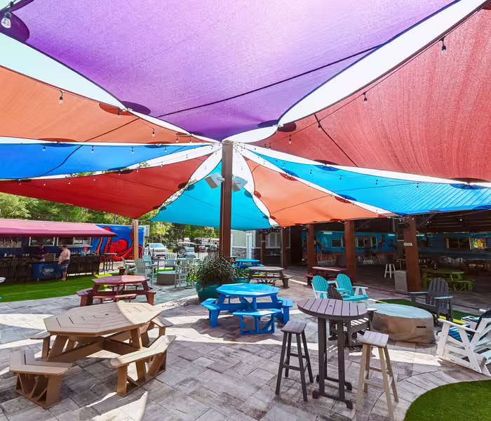 Colorful shade sails cover a paved patio with picnic tables and chairs at Splash RV Resort.