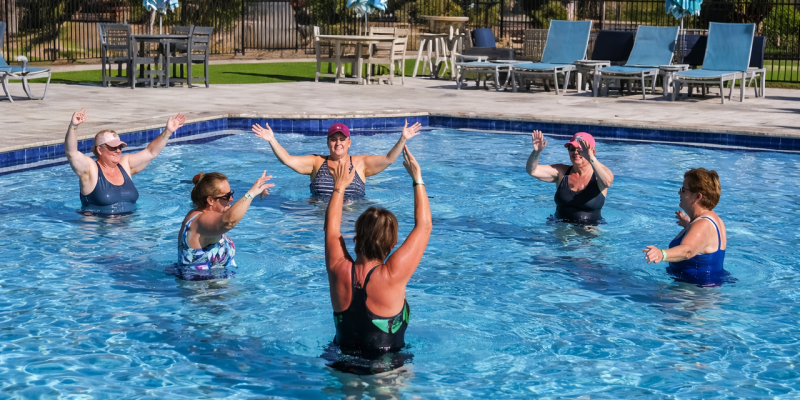Women doing water aerobics in the resort swimming pool at Splash RV Resort, surrounded by lounge chairs and umbrellas.